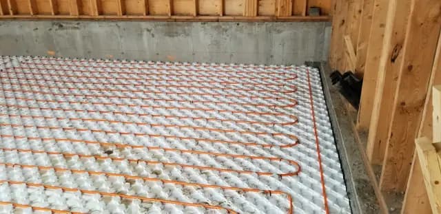 Orange and white radiant floor heating tubing is neatly arranged on a white insulation mat, with a concrete