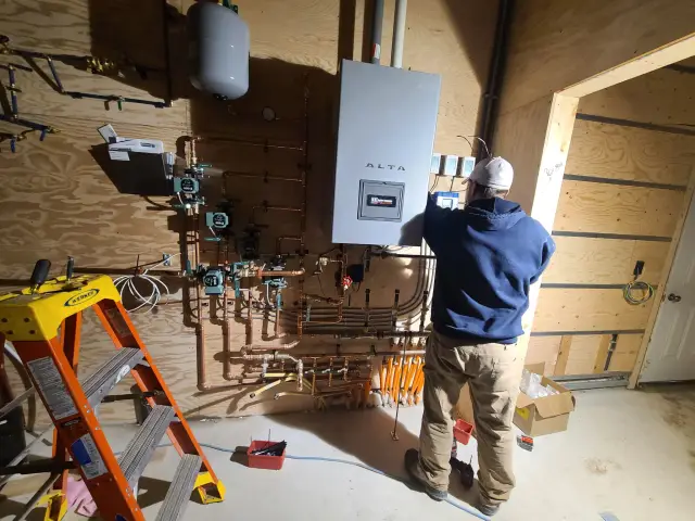 A is installing an Alta water heater onto a wall with various pipes and fittings. The setting appears