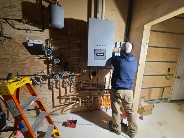 A is working on an unfinished wall with a large water heater labeled 'Alta'. The wall is covered