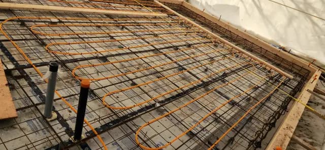 Orange radiant floor heating tubing is laid out on a white insulation layer with a wooden framework above