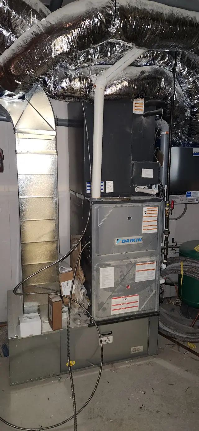 A Daikin furnace is installed in a home's utility room with ductwork and insulation visible above it. The