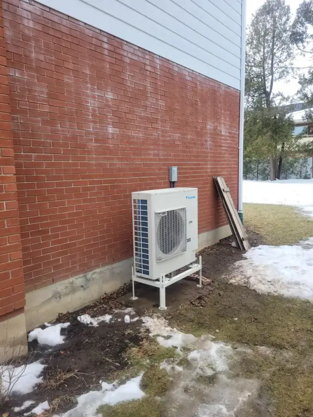 A new outdoor heat pump unit, presumably from the brand Gree, is installed against a brick wall. The