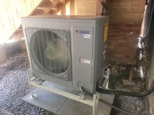A worker in a blue shirt and cap is carefully positioning a large, gray air conditioning unit with a circular fan into a wooden wall, highlighting the importan…