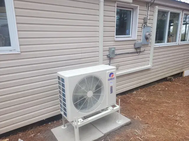 A worker in a blue shirt and cap is securing a large white air conditioning unit onto a concrete platform with a wrench.