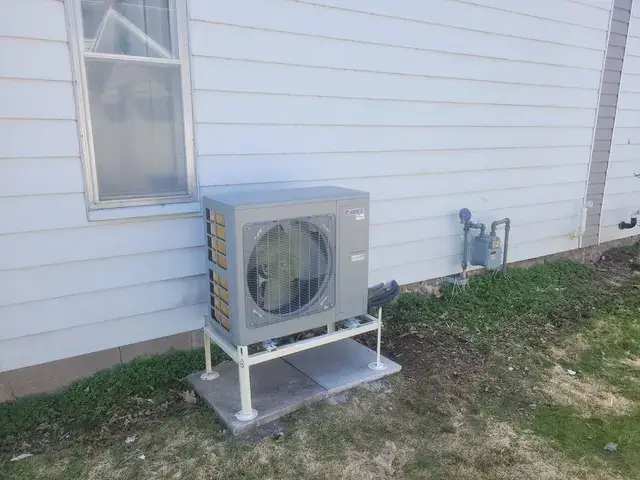 A large, gray outdoor heat pump is installed on a concrete platform outside a house. The heat pump