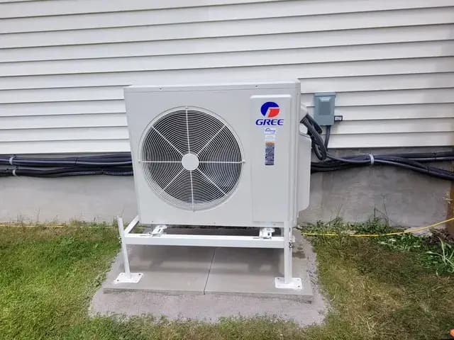 A technician, dressed in a blue shirt and dark pants, is closely examining a large white air conditioning unit with a circular fan, focusing on its components…