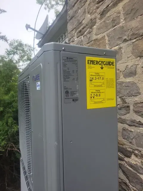 A large, gray outdoor heat pump unit with the brand 'Gree' visible is installed against a stone wall.