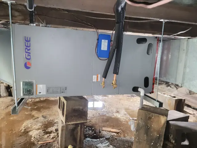 A new outdoor Gree heat pump is installed in a cluttered, unfinished space with a muddy ground and