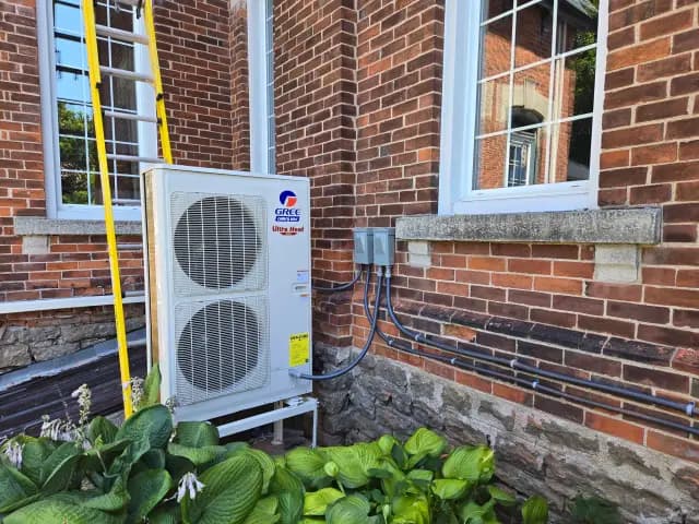 A large white outdoor unit with the Gree logo is installed against a brick wall with a ladder
