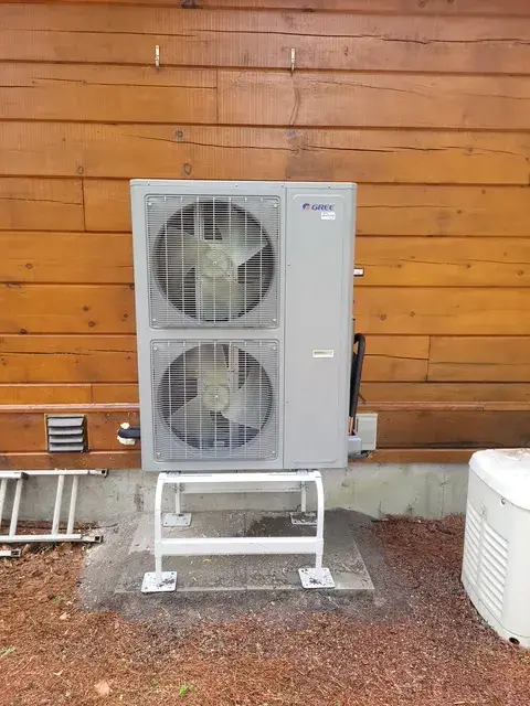 A large, gray outdoor heat pump with two fans is installed on a concrete slab against a wooden