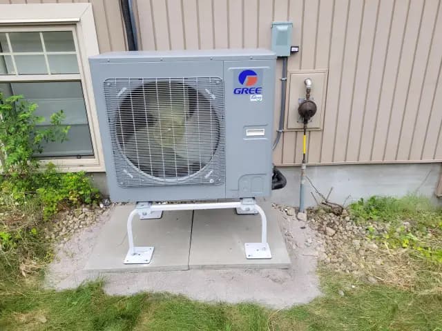 A large, gray outdoor heat pump with a circular fan is installed outside a house. The brand logo