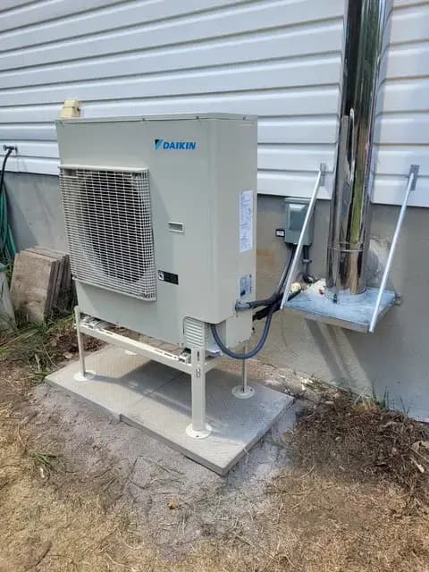 A Daikin heat pump unit is installed outside a house. It's a large, white box with a fan