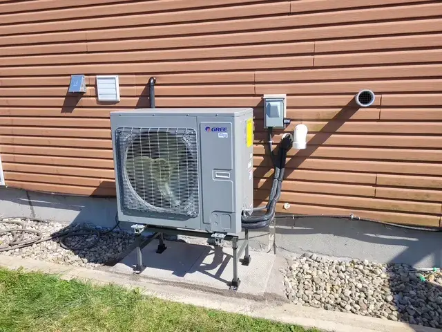 A large, gray outdoor heat pump with a fan is installed on a concrete pad against a house