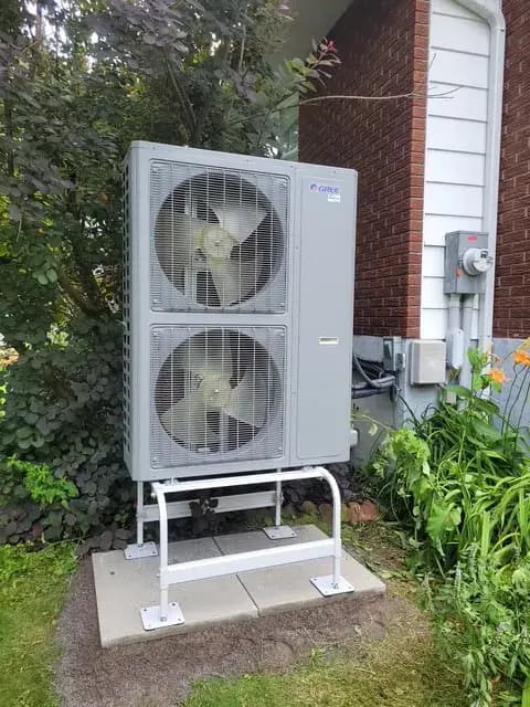 A large, gray outdoor heat pump with two fans is installed on a concrete platform. It is surrounded
