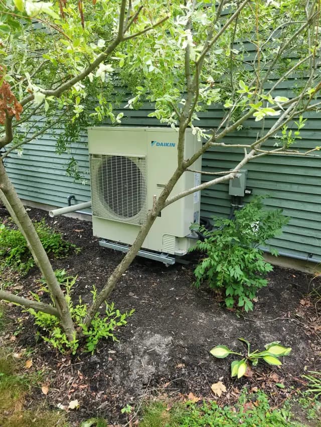 A Daikin heat pump unit is installed outside a house. It's a large, white, rectangular unit with a