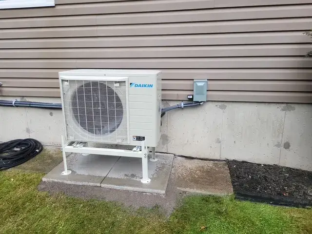 A Daikin heat pump unit is installed outside a house. It's a white, rectangular unit with a large