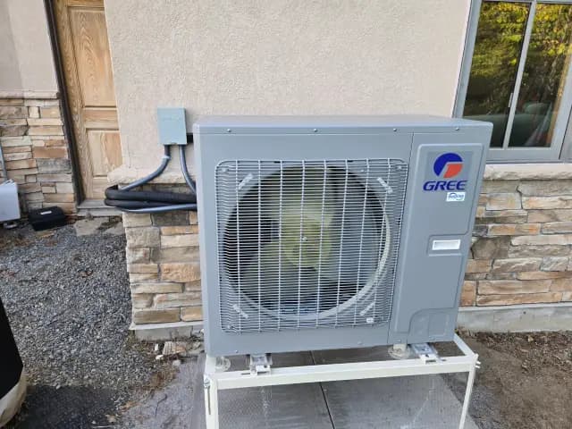 A large, gray outdoor heat pump with the Gree logo is installed on a stand outside a house.