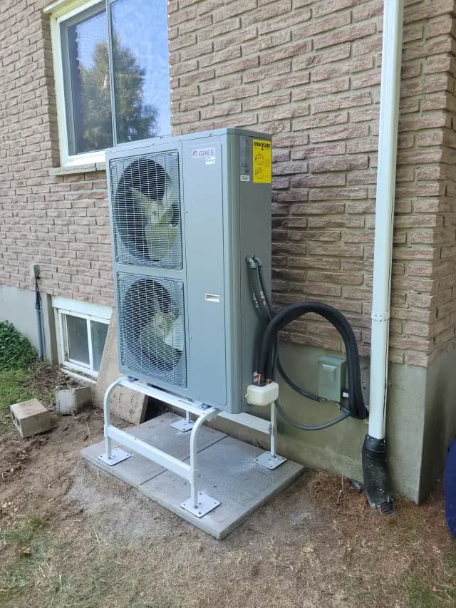 A large, gray outdoor heat pump with two fans is installed on a concrete platform outside a brick