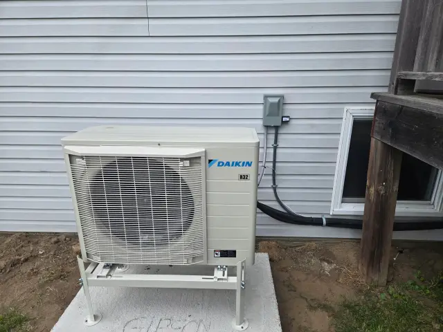 A Daikin heat pump unit is installed outside a house. It's a large, white, box-like structure with a
