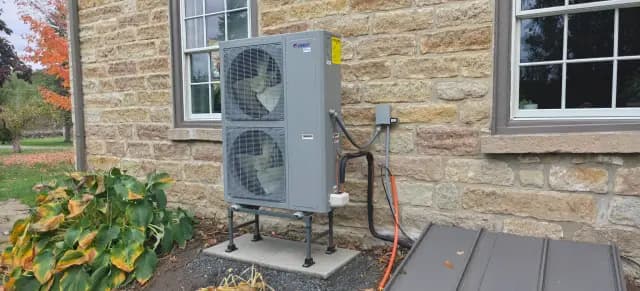 A large, gray outdoor heat pump with a fan is installed on the side of a stone house.