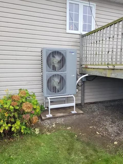 A large, grey outdoor heat pump with two fans is installed on the side of a house. It's
