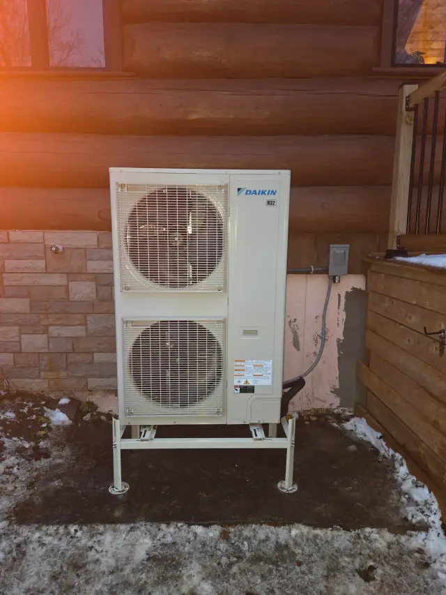 A Daikin heat pump unit is installed outside a house, with snow on the ground and a wooden