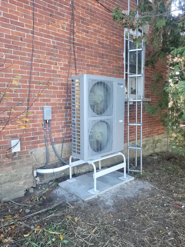 A large, grey outdoor heat pump unit with two large fans is installed on a concrete platform against