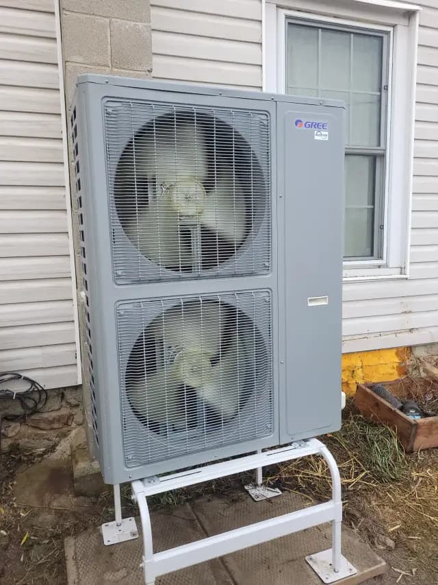 A large, gray outdoor heat pump with two large fans is installed outside a house. The brand 'Gree'