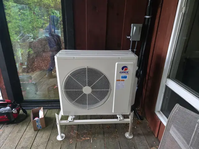 A large white outdoor unit with a circular fan is installed on a wooden deck. The brand 'Gree'