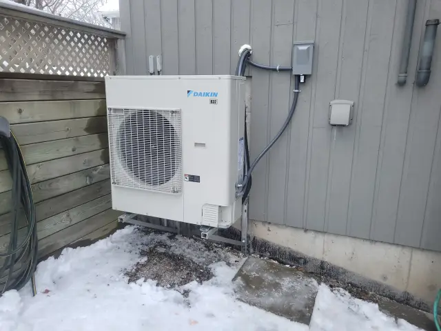 A Daikin heat pump is installed outside a house, with a large fan visible on the front. The