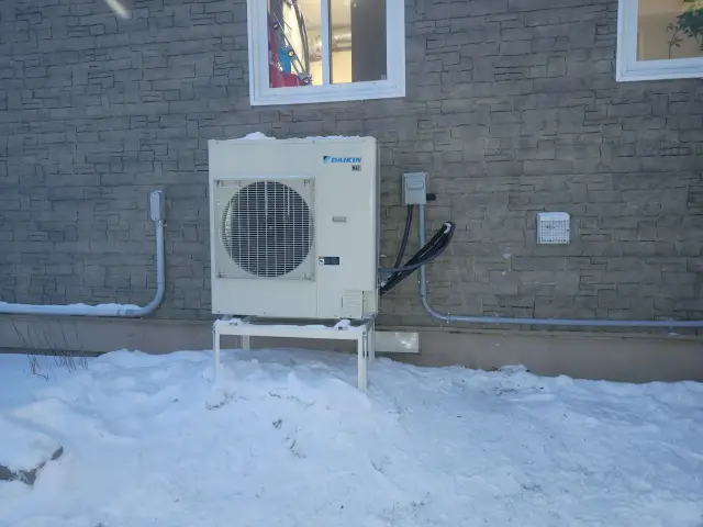 A Daikin heat pump unit is installed outside a building with snow on the ground. The unit is