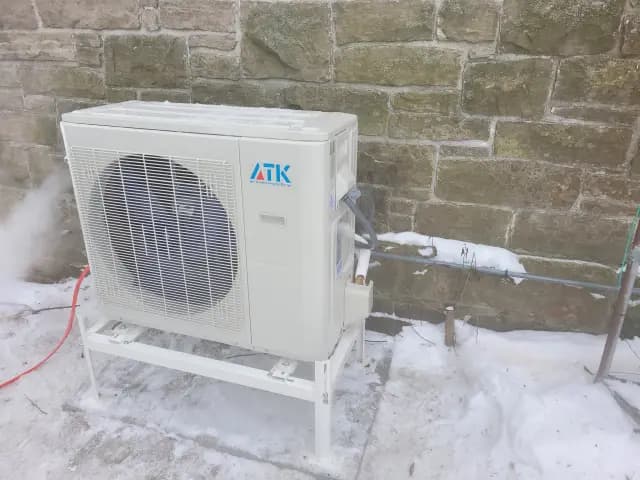 An outdoor Gree heat pump unit with a large fan, placed on a platform against a stone wall,