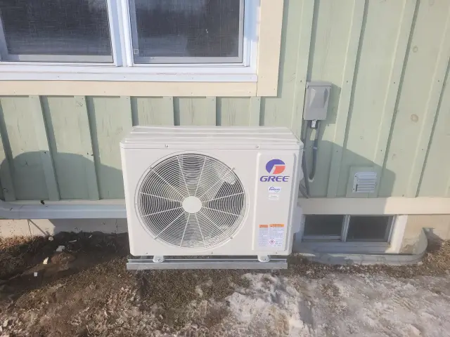 A large, white outdoor heat pump with the Gree logo is installed outside a house. The pump is