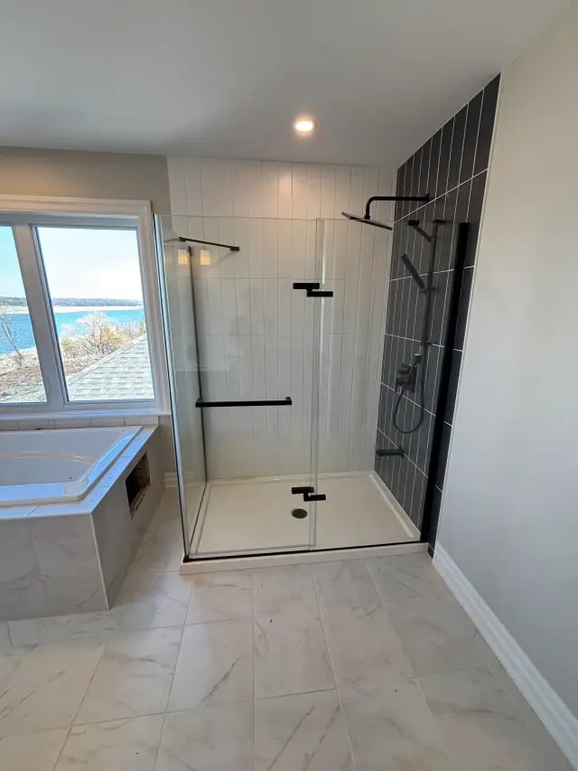 A spacious bathroom showcases a glass-enclosed walk-in shower with black fixtures, a white bathtub, and a large window