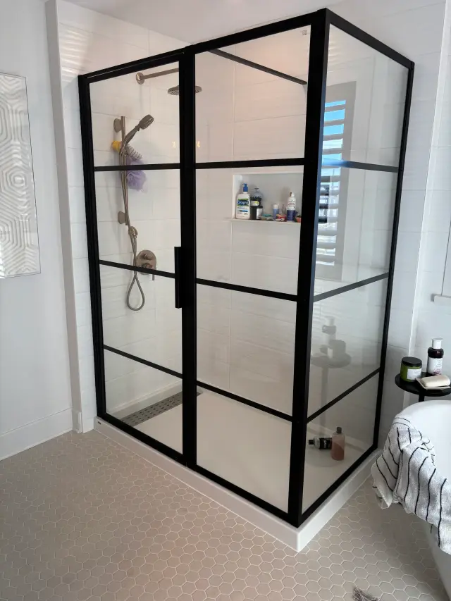 A black-framed glass shower enclosure with a showerhead, faucet, and visible shelving with toiletries