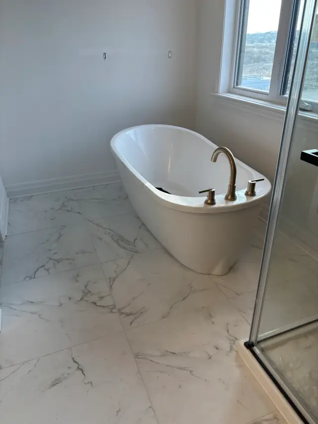 A white freestanding bathtub with a gold faucet is positioned against a white wall next to a window.