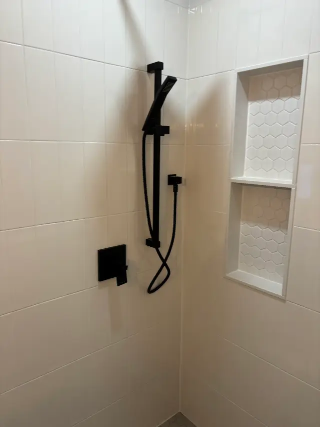 A close-up view of a modern shower setup, featuring a matte black overhead showerhead, a handheld showerhead, and