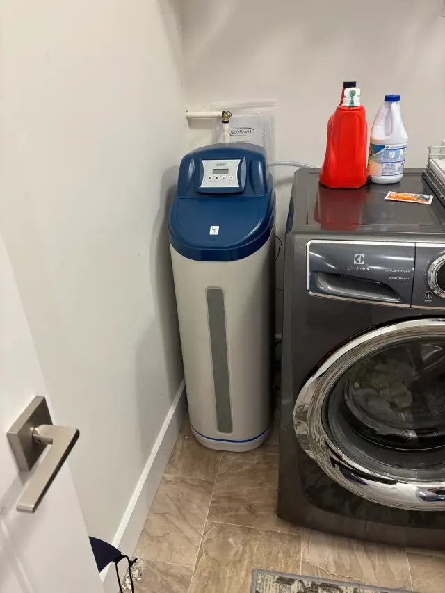 A water softener with a digital display is placed next to a front-loading washing machine. On the machine,