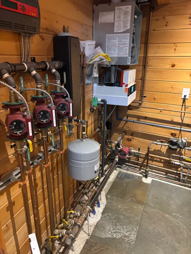 A digital control panel with red and black buttons is mounted on a wooden wall with various pipes