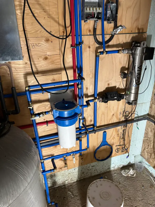 A close-up view of blue plumbing pipes with fittings and valves, including a white filter housing with a