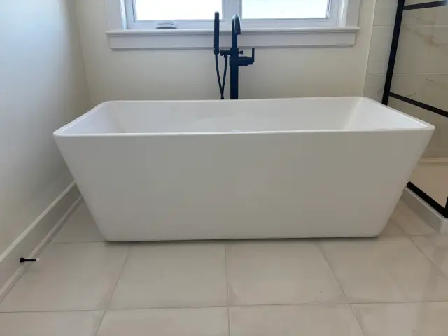 A white freestanding bathtub with a black faucet is placed in a bathroom with beige tiles. The tub