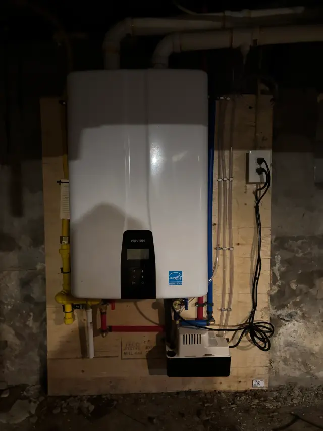 Navien tankless water heater with digital display, installed on a wooden board with various pipes and fittings around