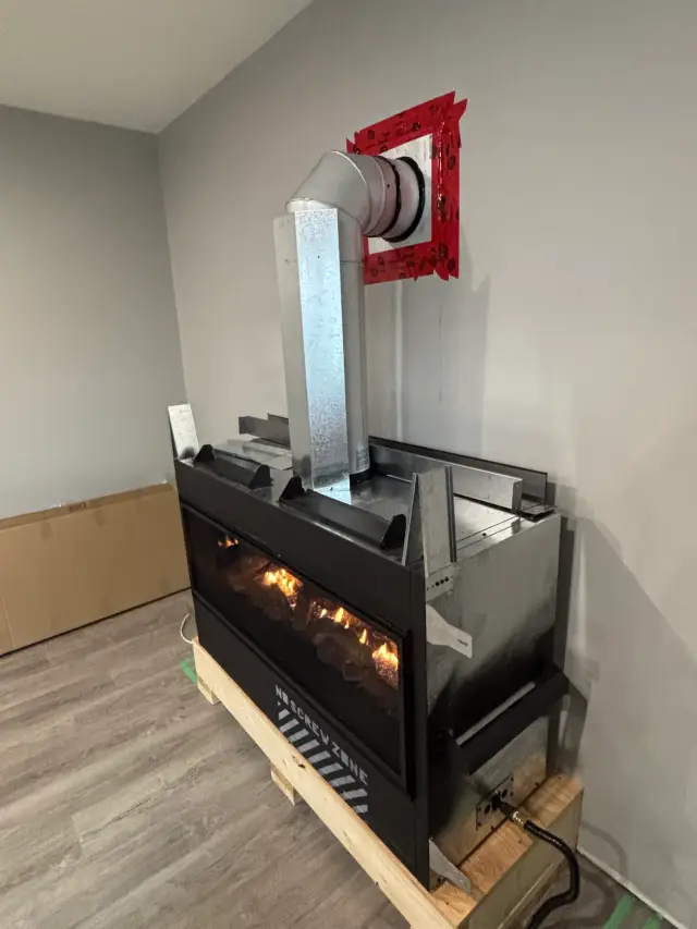 A modern gas fireplace with a reflective silver venting system is being installed in a room with a