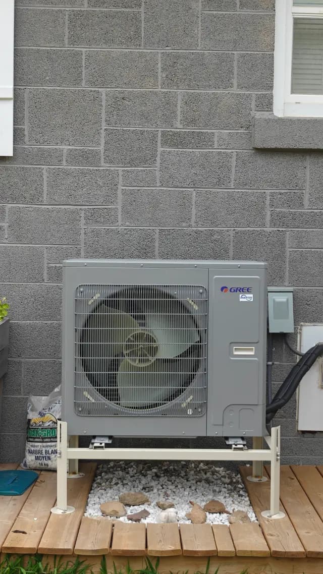 A new outdoor Gree heat pump is installed on a wooden platform against a gray brick wall. It's