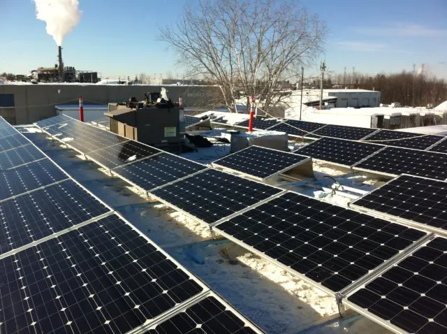 Solar panels on a roof with snow around them, with a factory in the background emitting smoke